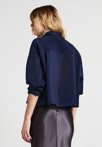 Donna con capelli biondi che indossa una blusa blu navy larga con maniche arrotolate e una gonna di raso scura, vista di spalle su uno sfondo neutro.