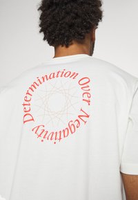 Valkoinen puuvillapaita lyhyillä hihoilla, jossa on punaisella tekstillä "Päättäväisyys Yli Negatiivisuuden" pyöreässä muodossa selässä.