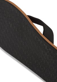 Musta kuminen flip-flop-pohja, jossa on teksturoitu pinta ja ruskea välikerros. Ominaisuudet kuusikulmainen kuvio, joka tarjoaa pidon. Minimalistinen muotoilu.