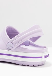 Ljuslila Crocs med ett band, med en strukturerad innersula, en vit sula med en lila rand och präglad logotyp på bandet.