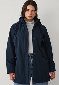 Kobieta ubrana w granatową kurtkę z kapturem zapinaną na zamek błyskawiczny i zatrzaski, pod którą ma biały golf i niebieskie dżinsy, stoi na jednolitym tle.