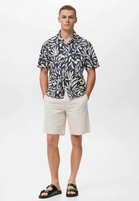 Camisa de manga corta en azul marino con un patrón de hojas blancas, combinada con pantalones cortos beige claros y sandalias negras con hebillas, de pie contra un fondo liso.