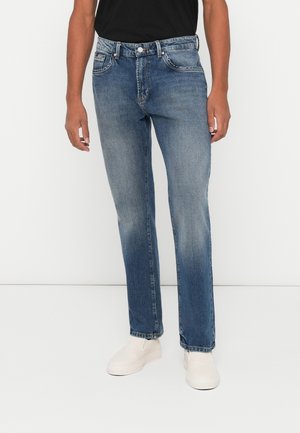 Personne portant un jean en denim bleu délavé, une chemise noire et des chaussures blanches sans lacets, se tenant devant un fond blanc uni.