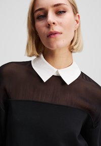 Haut noir avec un panneau transparent noir et un col blanc. Présente un design simple et une texture lisse, soulignant une esthétique moderne et minimaliste.