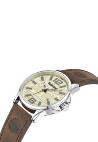 Reloj analógico redondo con un dial texturizado en beige, números en tono plateado y caja de acero inoxidable. Correa de cuero marrón con detalle de logo en relieve.