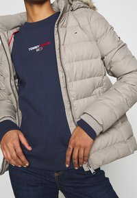 Osoba w jasnobeżowej kurtce puchowej i granatowej bluzie z logo "TOMMY JEANS", z rękami spoczywającymi na biodrach, pokazując tatuażowane palce.