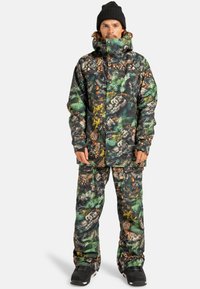 Mężczyzna ubrany w pasującą kurtkę zimową i spodnie w kamuflażowy wzór z motywem szyszek i liści, czarną czapkę beanie i czarne buty.