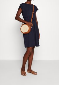 Granatowa sukienka z krótkimi, falbaniastymi rękawami; z wcięciem w talii. Okrąła torba przekłada na ramię z plecionego słomy z jasnobrązowym skórzanym paskiem. Jasnobrązowe płaskie sandały.