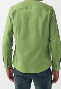 Grünes Langarmhemd mit Button-Down-Kragen, das eine glatte Textur und seitliche Schlitze aufweist, kombiniert mit dunklen Jeans.