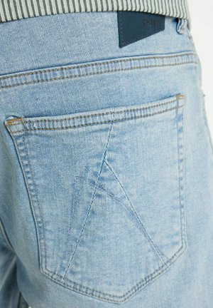 Achterzak van lichtblauwe spijkerbroek met dubbele stiknaad en zwart merkje boven de zak op de tailleband van een gestreept shirt.