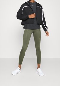 Veste noire courte avec accents blancs, t-shirt gris, leggings verts avec poches latérales et chaussures de sport blanches.