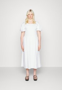 Vestido branco com mangas bufantes, decote quadrado e corpete franzido. Desce até a metade da panturrilha. Usado com sandálias de plataforma pretas.