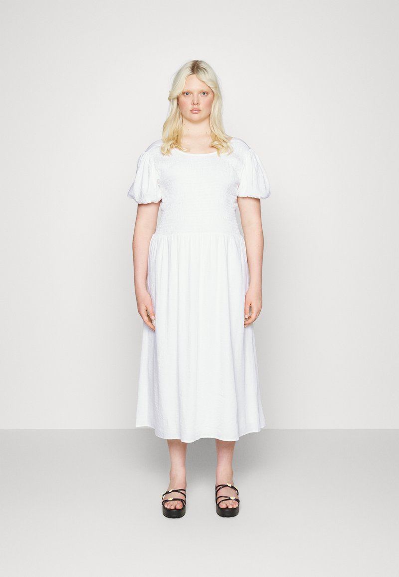 Vestido branco com mangas bufantes, decote quadrado e corpete franzido. Desce até a metade da panturrilha. Usado com sandálias de plataforma pretas.