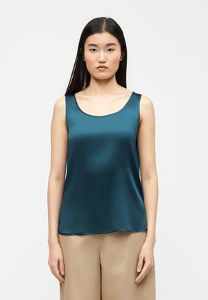 Femme aux longs cheveux noirs portant un haut en satin bleu sarcelle sans manches et un pantalon ample beige sur un fond blanc.