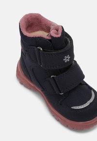 Stivale in tessuto blu navy con interno in fleece rosa, cinturini in velcro e suola in gomma. Presenta un piccolo accento a forma di fiocco di neve sul cinturino.