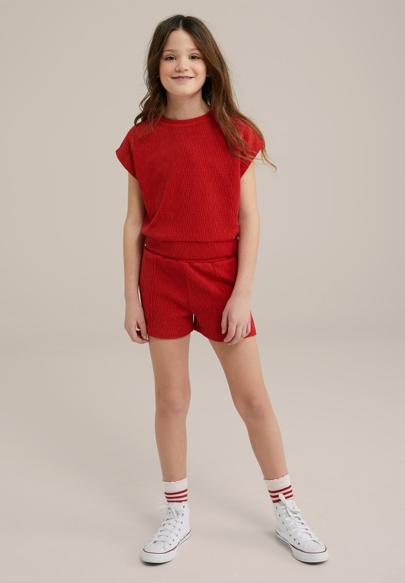 Piros bordázott, rövid ujjú felső, hozzá illő sorttal párosítva. Fehér, magas szárú sportcipők piros csíkozással és térdig érő zoknikkal. Egyszerű háttér.