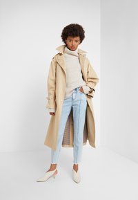 Beige trench coat with wide lapels, a textured cream turtleneck, light blue jeans, and cream heeled shoes, featuring subtle patterns inside.