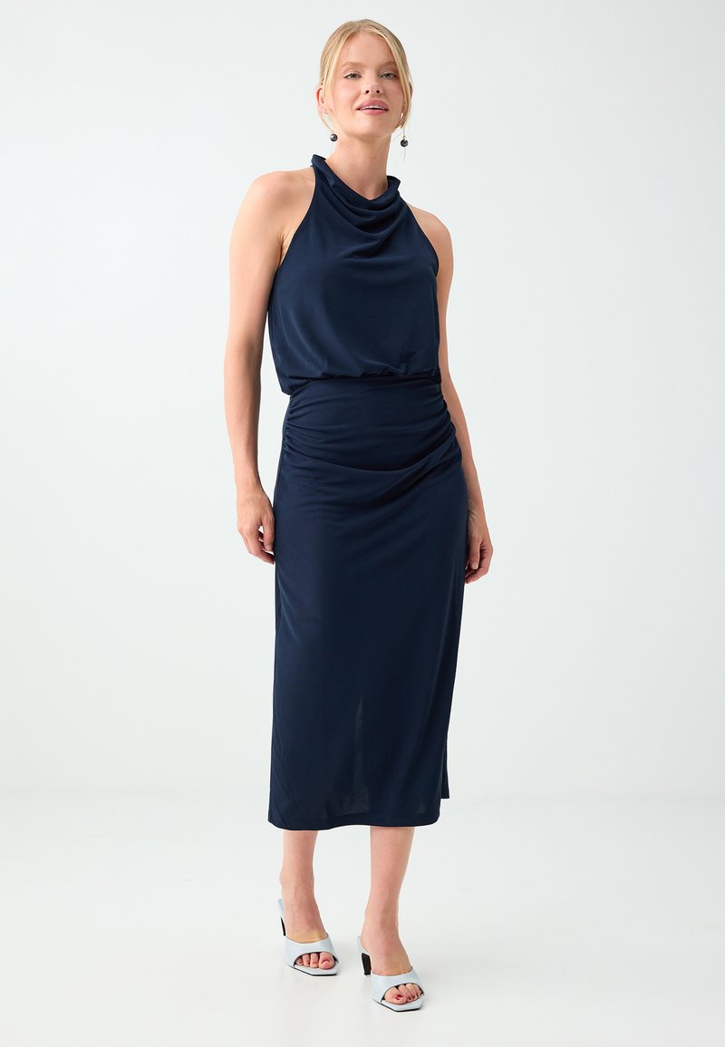 Femme aux cheveux blonds porte une robe bleu marine sans manches à col bénitier et des talons blancs ouverts à l'avant, debout devant un fond clair uni.