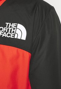 Červeno-černá bunda s hladkou texturou, geometrickým blokováním barev a výrazným bílým logem na horní levé straně.