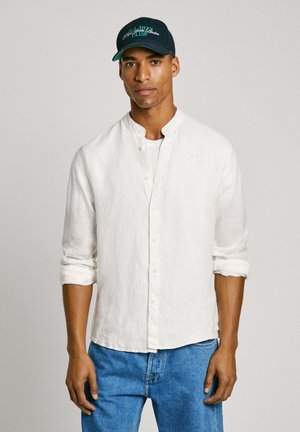 Camicia bianca in lino con maniche arrotolate, abbinata a jeans di denim blu e un cappellino navy con testo verde.