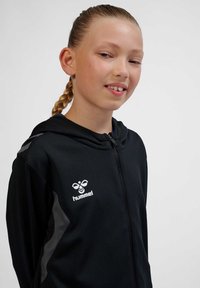 Ung pige med flettet hår iført en sort Hummel lynlåshættetrøje, der smiler let mod en ensartet lys baggrund.