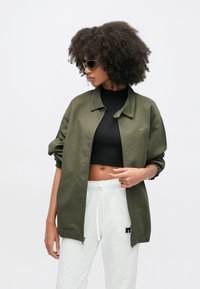 Ζακέτα ελιάς με φερμουάρ, γιακά, μανίκια με σούρες και διακριτικό λογότυπο. Συνδυασμένη με μαύρο τοπ κ�αι ανοιχτό γκρι φόρμες.