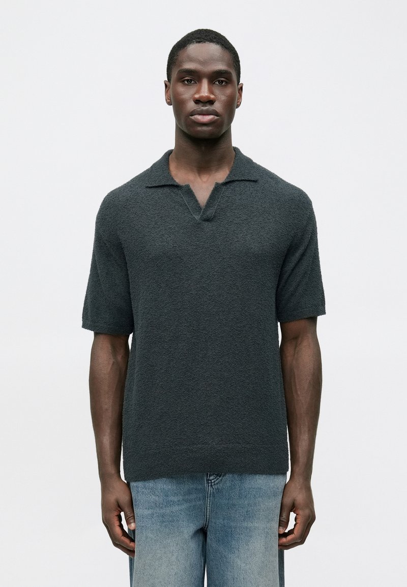 Σκούρο γκρι πλεκτό πόλο μπλουζάκι με κοντό μανίκι, υφή και σχέδιο με V-λαιμόκοψη. Συνδυάζεται με ανοιχτό μπλε τζιν παντελόνι.