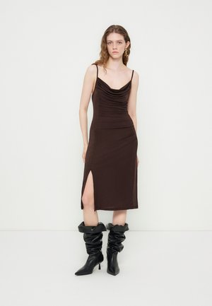 Jeune femme portant une robe midi marron sans manches avec une fente latérale et des bottines noires à talons souples, debout contre un mur blanc uni.