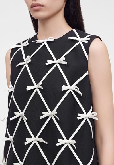 Femme portant une robe noire sans manches avec un motif en treillis blanc formé de bretelles croisées nouées en petits nœuds.