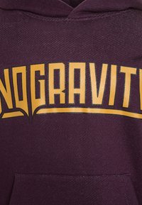 Fialová mikina s velkým žlutým grafickým nápisem "NOGRAVITY" v stylizovaném písmu a přední kapsou. Viditelná měkká textura tkaniny.
