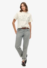 Hvid grafisk t-shirt med "SUPER DRY" tryk, parret med stribede blå og hvide bukser og brunt bælte. Sorte træskor afslutter outfittet.