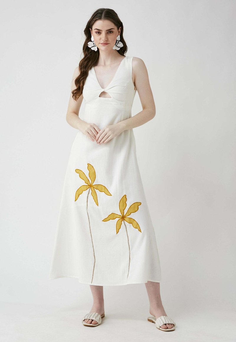 Weißes Maxikleid aus Leinen mit V-Ausschnitt und einem Cutout vorne. Das Rockteil ist mit gelben gestickten Palmen versehen. Die Pantoletten haben ein gerafftes Design.