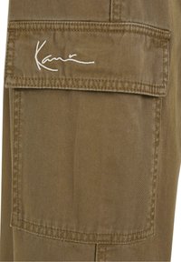 Poche cargo en tissu marron avec rabat et signature blanche brodée dans le coin supérieur gauche du rabat.