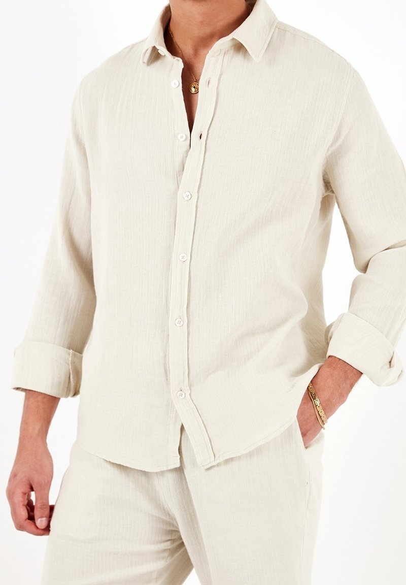 Camicia a maniche lunghe beige chiaro con bottoni, realizzata in tessuto testurizzato e dotata di colletto e polsini arrotolati, presenta bottoni bianchi sul davanti.