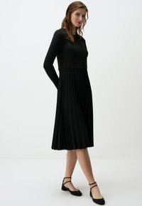 Robe noire à manches longues avec un corsage ajusté passant à une jupe plissée. Inclus des chaussures noires à talon plat avec des brides de cheville.