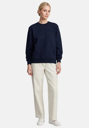 Mujer con sudadera azul marino, pantalones rectos beige claro y zapatillas blancas, de pie frente a un fondo liso.