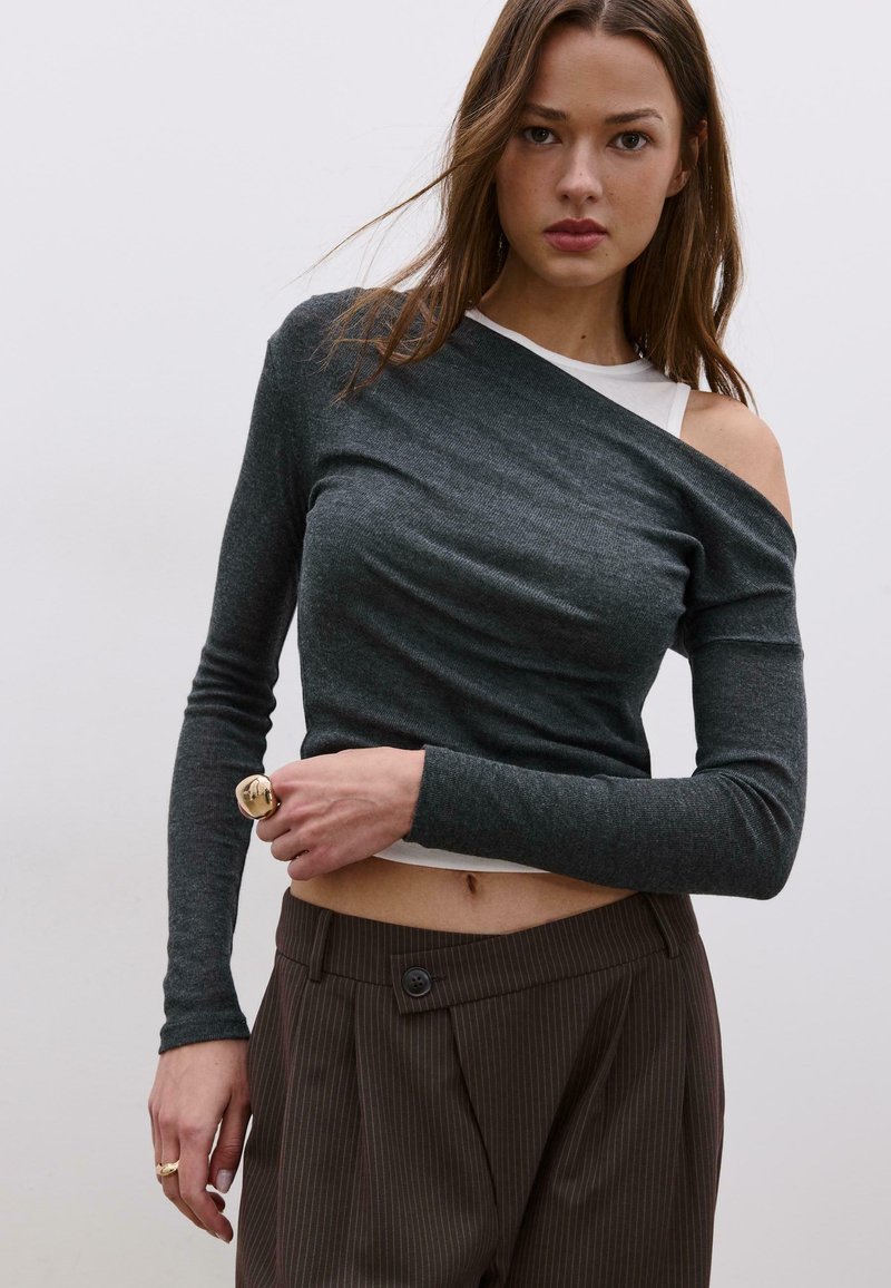 Donna che indossa una maglia a maniche lunghe grigio scuro scivolata sulle spalle sopra una canottiera bianca, pantaloni marroni a righe sottili e anelli d'oro sulla mano sinistra.