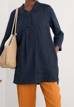 Personne portant une tunique bleu foncé à manches longues et un pantalon orange tenant un sac fourre-tout tissé beige avec des poignées en corde.