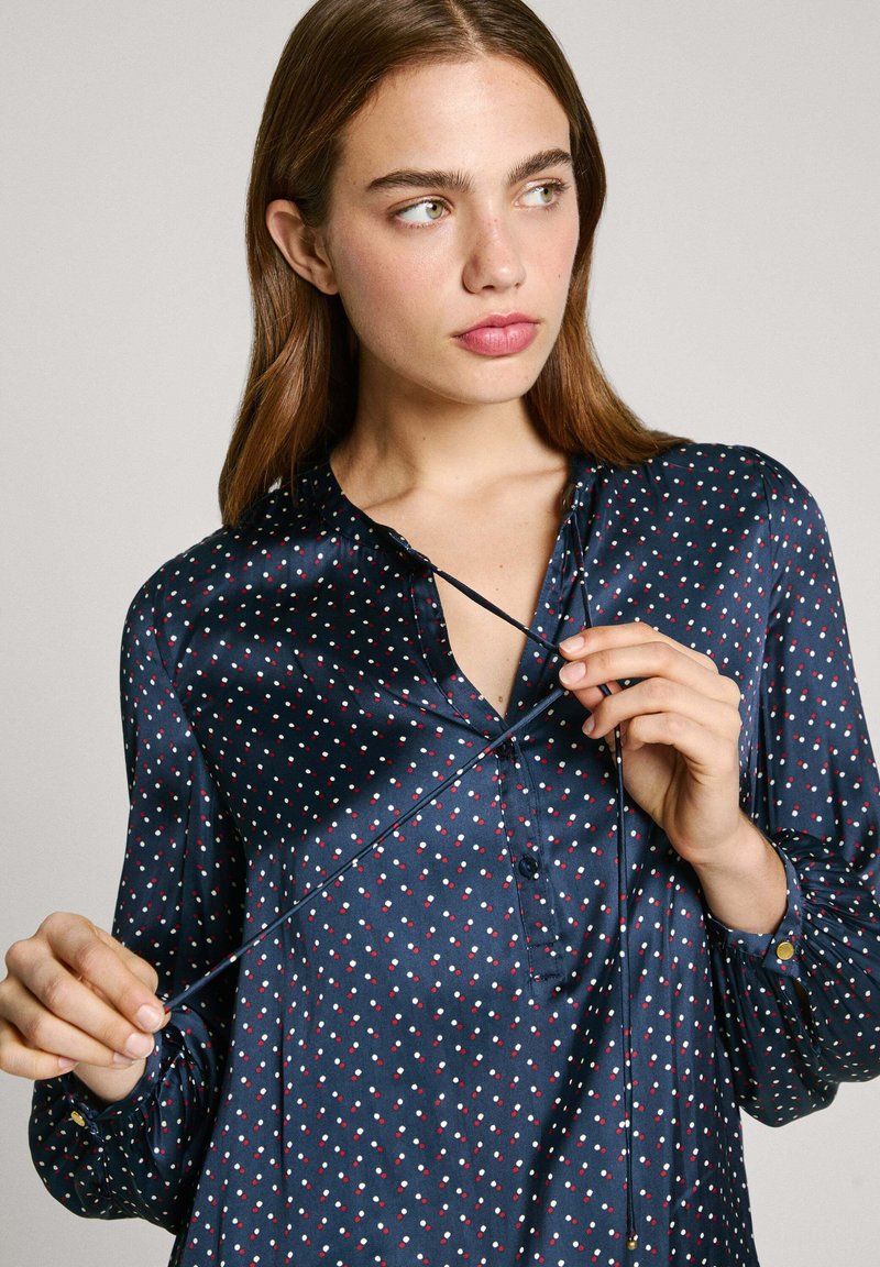 Blusa de marina en satén con lunares blancos y rojos, con botones en la parte delantera y un puño ligeramente fruncido. Presenta un corte holgado y cuello.