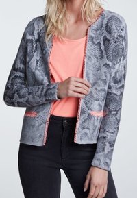 Cardigan tricot à imprimé serpent gris avec des accents orange. Il présente une coupe décontractée et un design ouvert à l'avant, associé à un t-shirt rose en dessous.