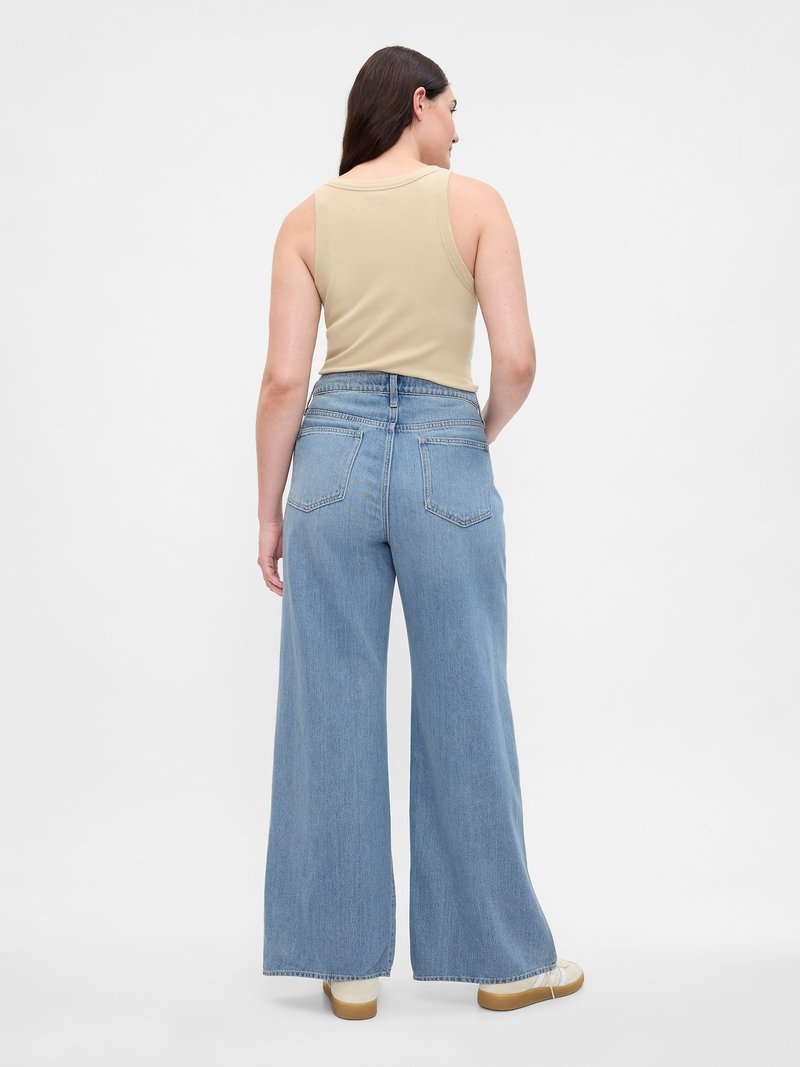 Frau, die mit dem Rücken zu sehen ist, trägt ein beige ärmelloses Top, hochtaillierte, weite blaue Jeans und weiße Turnschuhe vor einem schlichten weißen Hintergrund.