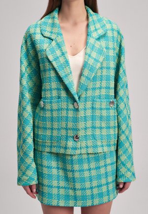 Vrouw draagt een turquoise en lichtgroene geruite jas en bijpassende rok met zilveren knopen over een wit geribd topje.