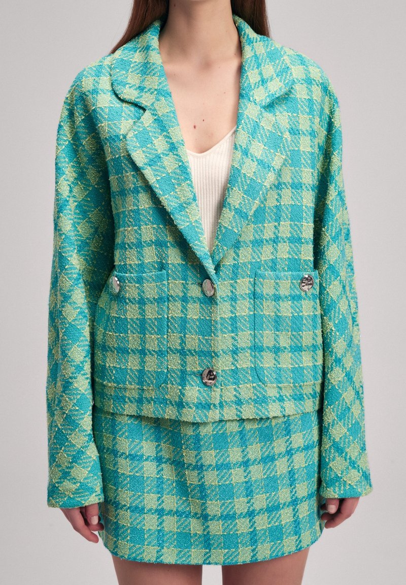 Femme portant une veste à carreaux turquoise et vert clair et une jupe assortie avec des boutons argentés sur un haut blanc côtelé.