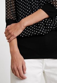 Schwarzes Oberteil mit durchsichtigen Ärmeln mit Polka-Dots, lockerer Passform und rundem Ausschnitt. Ausgestattet mit einem einfarbig schwarzen Oberteil und weißem Punktmuster.