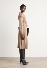 Kleid in Beige mit langen Ärmeln, tailliert mit einem Gürtel, goldenen Knöpfen und einem ausgestellt Rock. Kombiniert mit schwarzen, oberschenkelhohen Stiletto-Stiefeln und einer schwarzen Tasche.