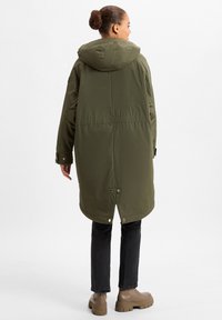 Cappotto lungo verde oliva con cappuccio, presenta una vita accentuata, bottoni a pressione e spacchi laterali. Abbinato a jeans neri e stivali marroni.