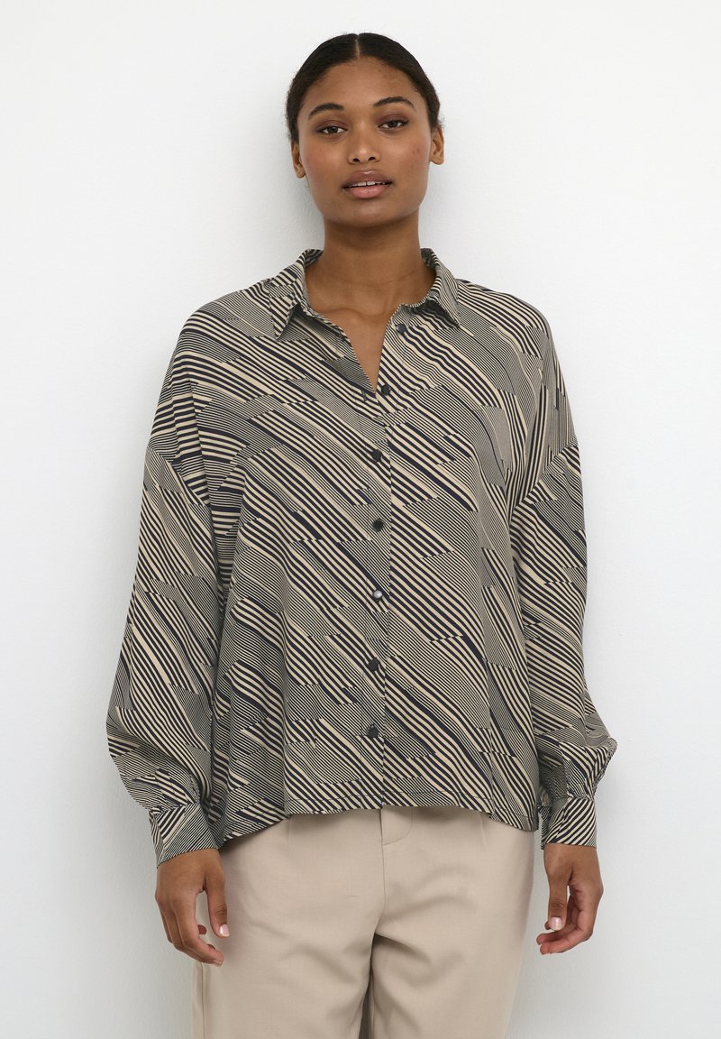 Frau in beigen Hosen und einem übergroßen, beige-schwarz gemusterten Hemd, das gegen einen weißen Hintergrund steht.