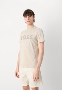 Beige pamut póló, amelyen egy sötétebb árnyalatú "BOSS" logó található, kerek nyakkivágással, világos színű rövidnadrággal párosítva.