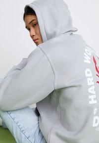 Ljusgrå hoodie med en framficka i känguru-design, med vit och röd grafisk text på baksidan. Tyget verkar mjukt och texturerat.