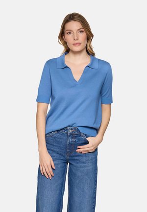 Mujer con una camisa polo de manga corta azul claro metida dentro de unos vaqueros de talle alto azules, de pie con una mano en el bolsillo.